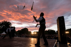 Foto eines Mannes, der auf der Bühne steht und jongliert. Die Sonne geht gerade unter und das Publikum ist im Hintergrund zu sehen.