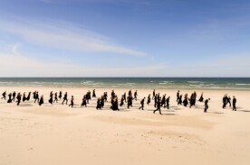 Fotografie eines Strandes, an dem mehere schwar gekleidete Personen entlang laufen.