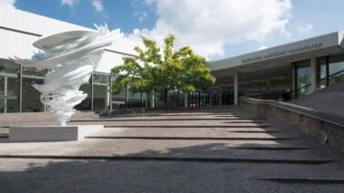 Eingangsbereich des Sprengel Museums Hannover. Auf der linken Seite ist ein weißes Kunstwerk und ein Baum zu sehen. Der Himmel ist blau mit ein paar Wolken und die Sonne scheint.